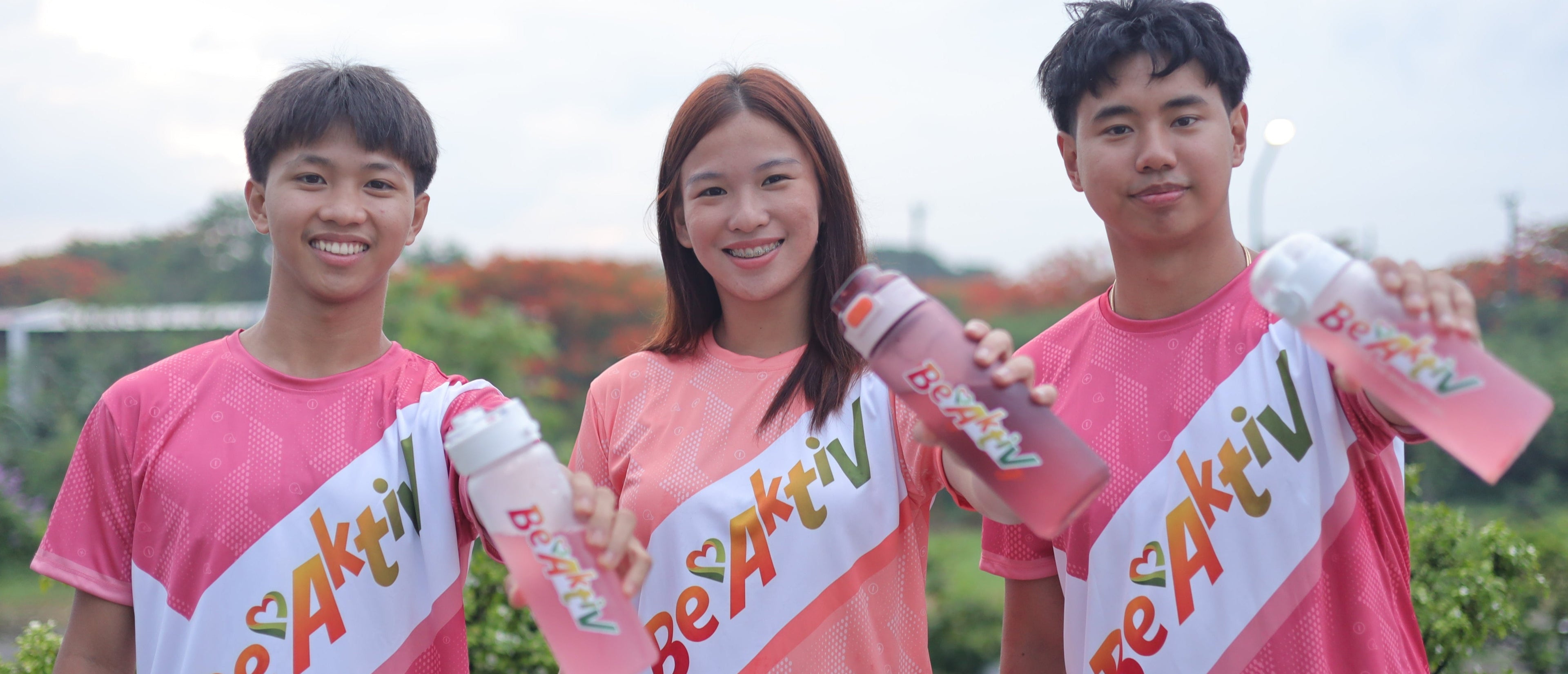 Three people wearing pink shirts with 'Be Aktiv' branding, holding bottles against a blurred outdoor background.